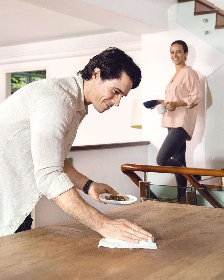 Man cleaning a table, woman smiling and observing with a cup and bowl in hand (Photo)